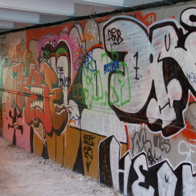 A wall covered with many instances of overlapping graffiti, abstract word tags and stylised faces. Red and organce dominates with some green and pink sections, with strong black and white lines and some concrete still visible beneath.