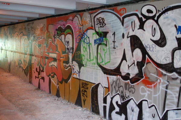 A wall covered with many instances of overlapping graffiti, abstract word tags and stylised faces. Red and organce dominates with some green and pink sections, with strong black and white lines and some concrete still visible beneath.