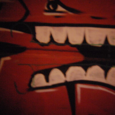 A picture of a stylised spray-painted square orange face with large white teeth, in an angry expression. Graffiti taken with a Holga plastic lens.