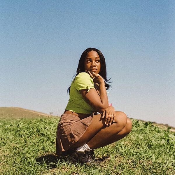 Rachel wearing a yellow top and brown skirt, crouched on a grassy hill with a hazy blue sky in the background.