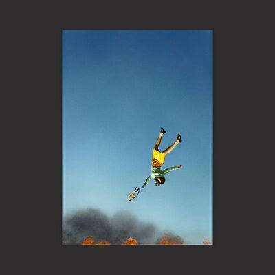 Artwork for Spitting Off the Edge of the World features a woman in a yellow pencil skirt and green jumper falling head first into a raging fire.