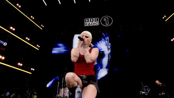 Cassyette kneels on stage at BBC Radio 1's Big Weekend. In the background the band are visible, with a big screen showing Cassyette behind them.